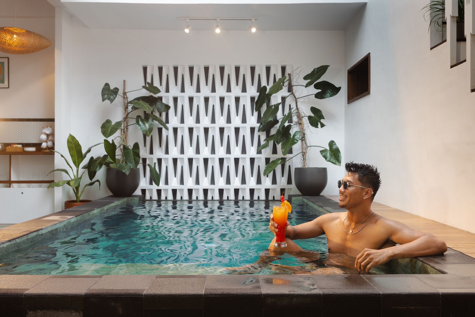 Man in sunglasses in a pool, holding a cocktail.  Plants and patterned wall in a white-walled room.