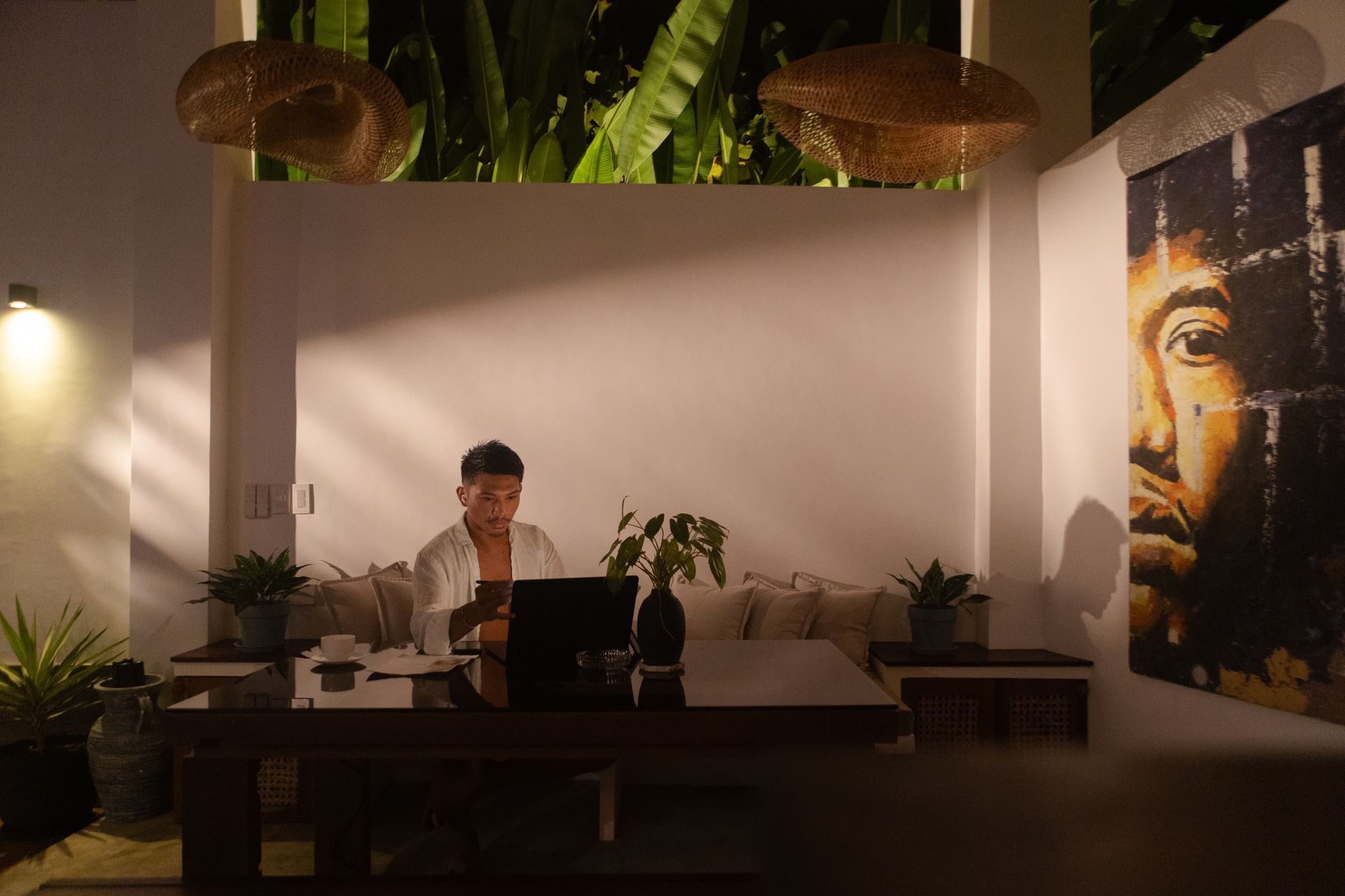 Man working on laptop at a desk, dimly lit outdoor setting, with art and plants.
