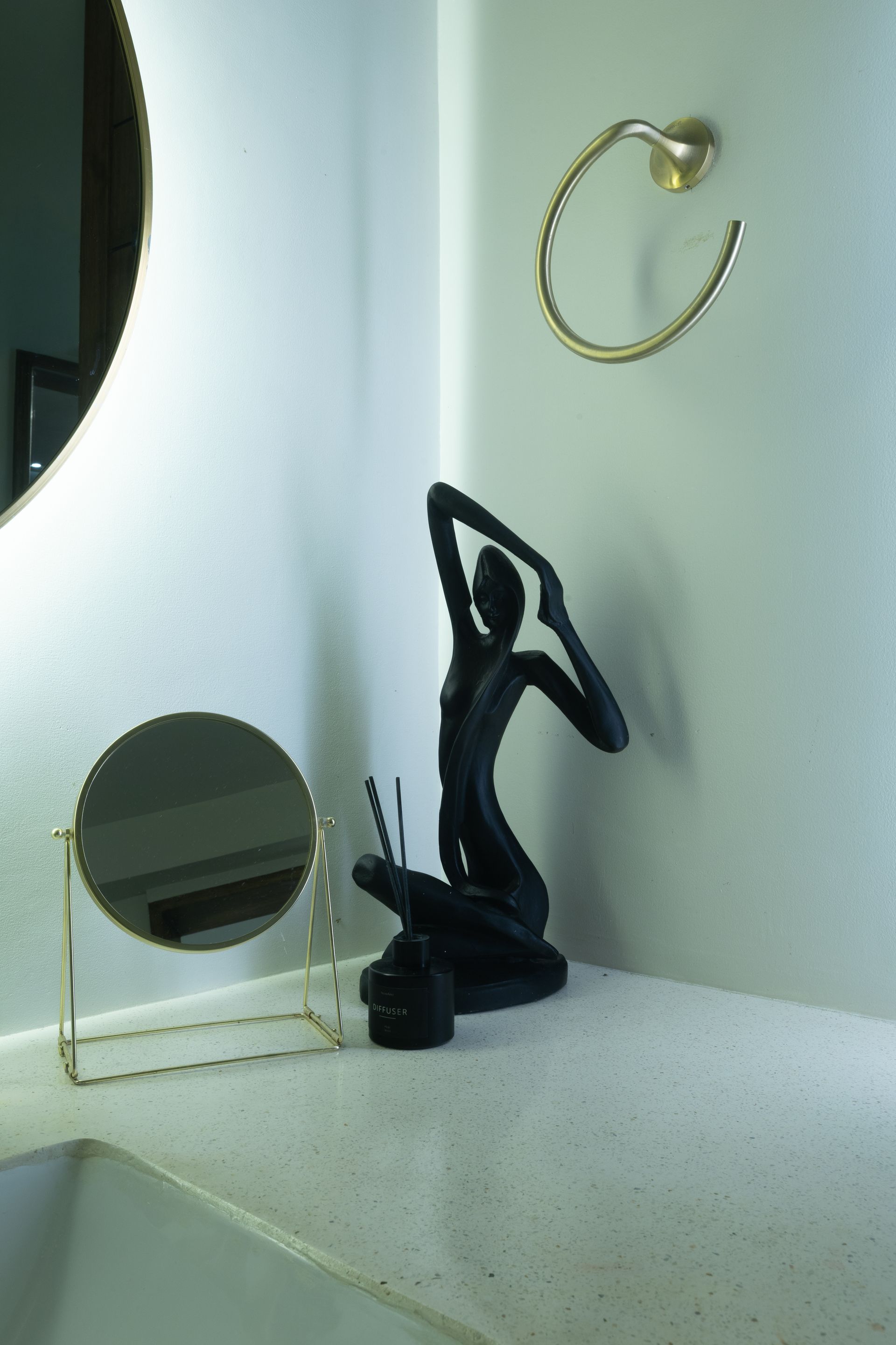 A bathroom corner with a black sculpture, gold mirror, towel ring, and a small mirror.