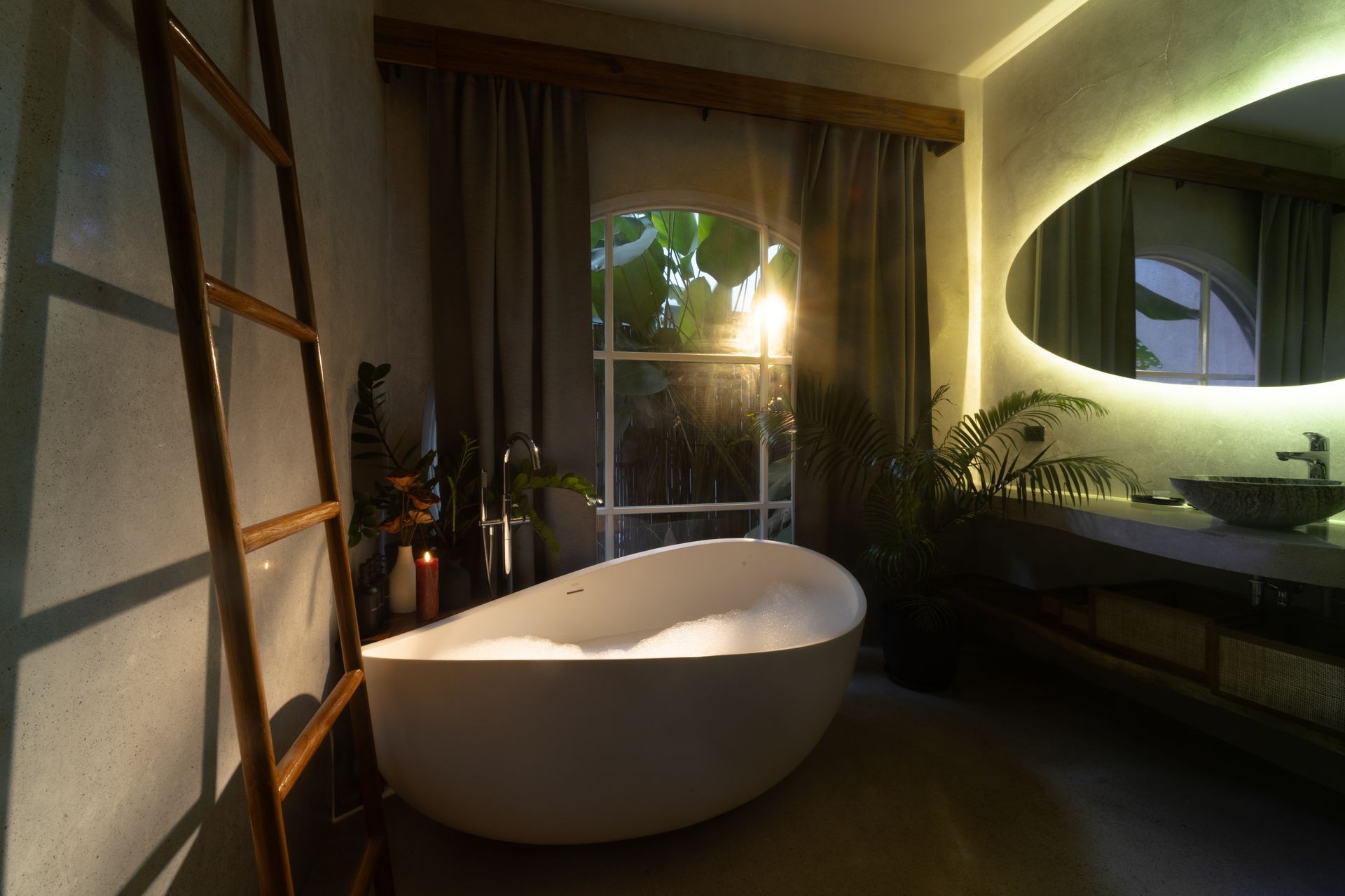 Cozy bathroom with a freestanding tub, ladder, and oval mirror with backlighting. Light streams in from the window.