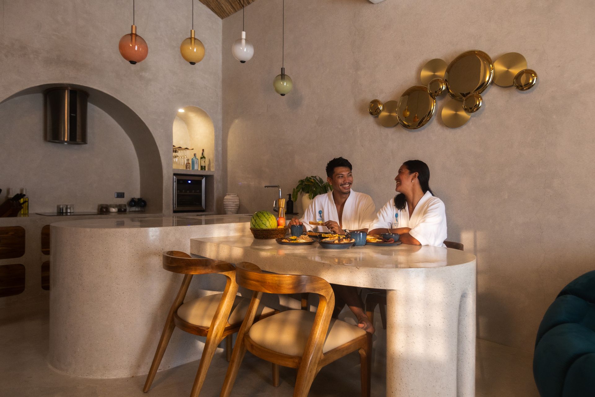Couple in robes laughing at a table in a bright room with pendant lights and decorative wall art.