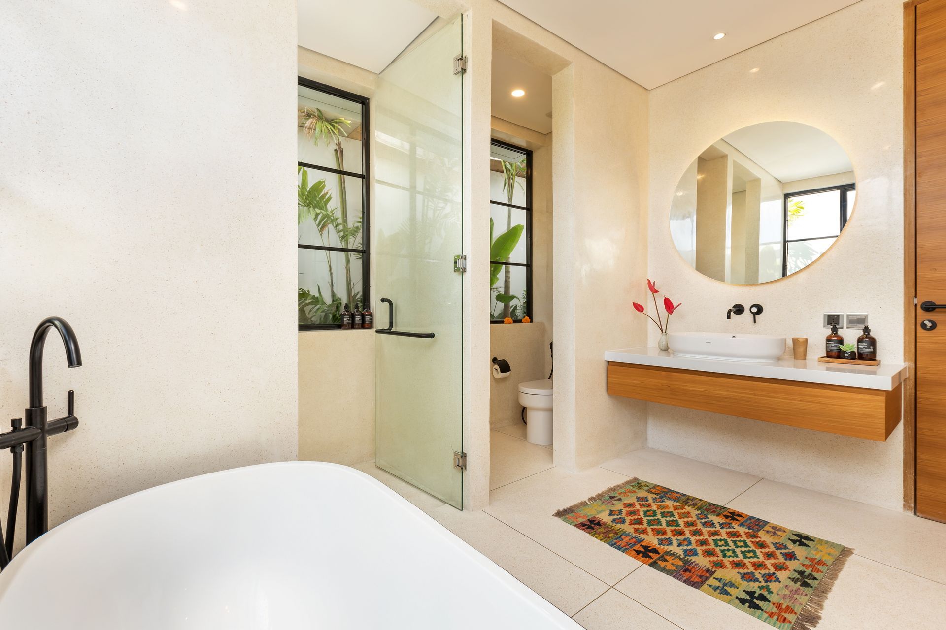 Modern bathroom with bathtub, shower, and vanity. Neutral tones, a round mirror, and a colorful rug add warmth.