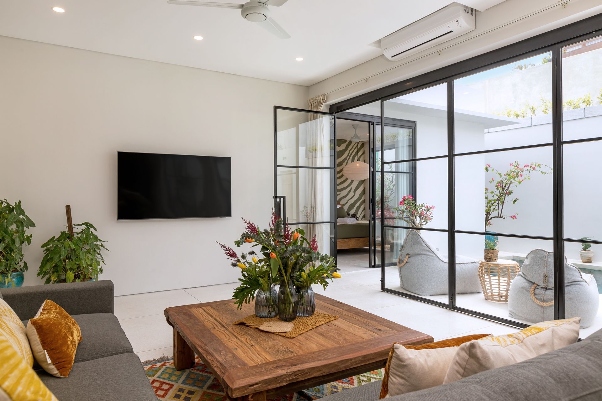 Living room with glass doors, sofa, TV, wooden table, and plants.