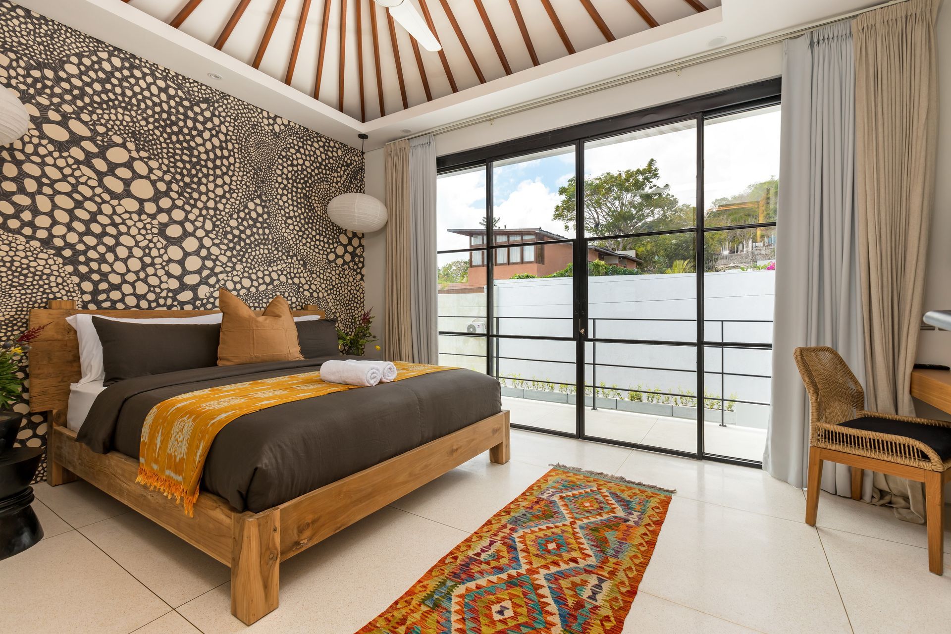 Bedroom with patterned wallpaper, large window, wooden bed, and colorful rug.