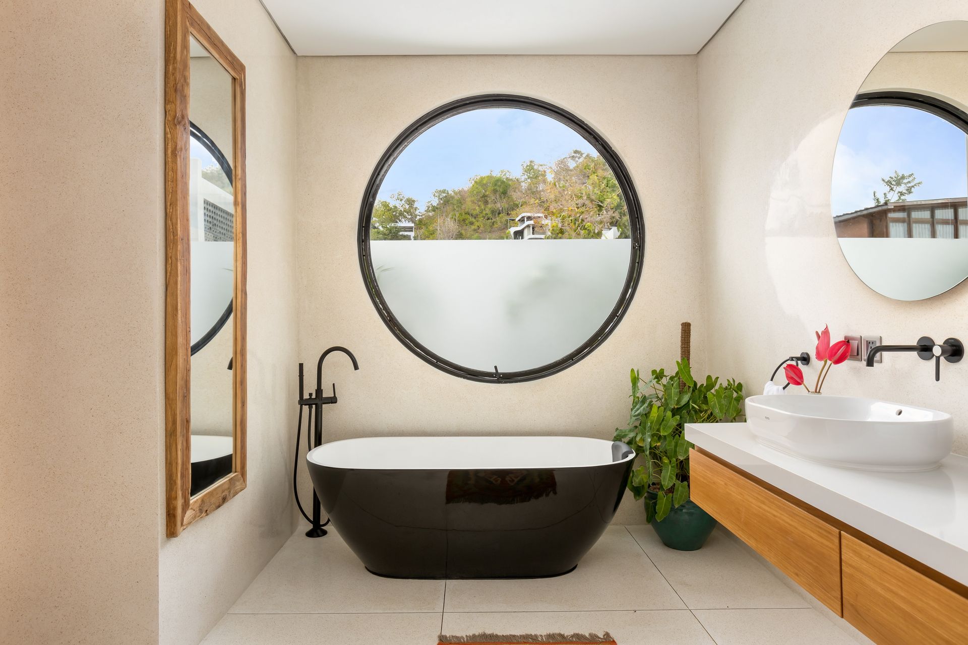 Bathroom with black tub under circular window, round mirror, and wooden vanity.