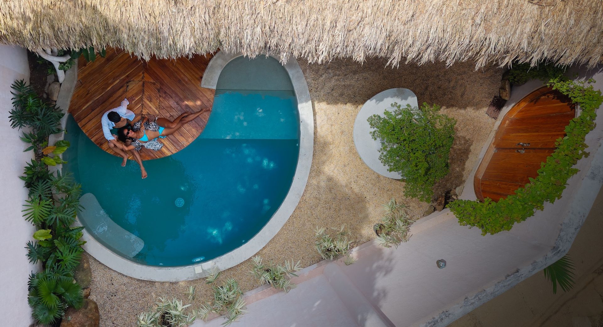 Overhead view of a small pool with a wooden deck, two people, and a thatched roof.