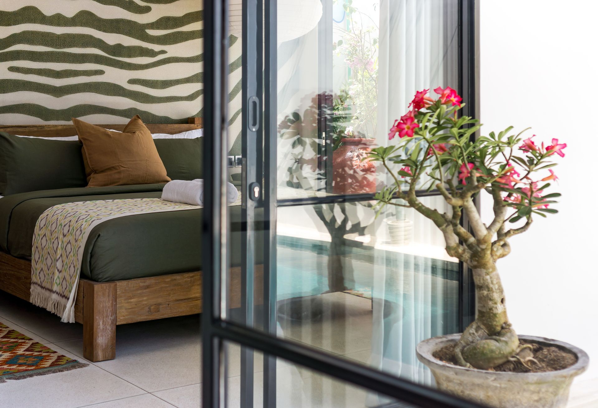 Bedroom interior with olive green bedding, a wooden bed frame, and a potted plant outside a glass door.