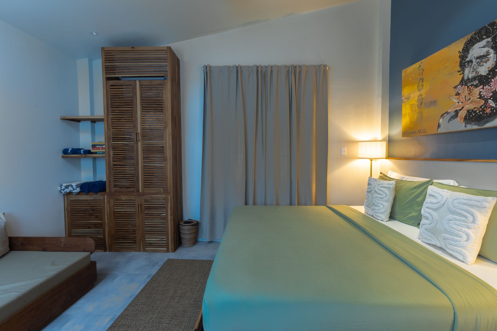 Bedroom with bed, wooden wardrobe, and artwork. Green bedspread, neutral curtains, and blue accent wall.