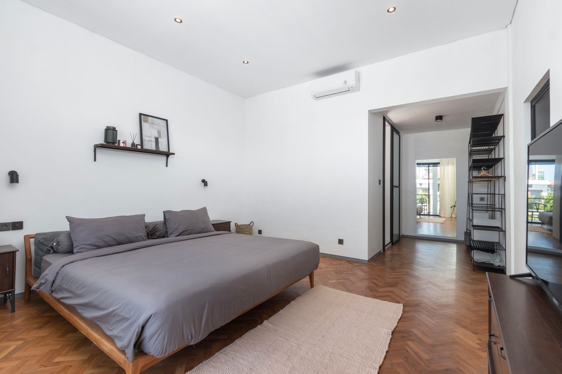 Bedroom with a bed, rug, shelving, and access to a balcony; wooden floor, white walls, gray bedding.