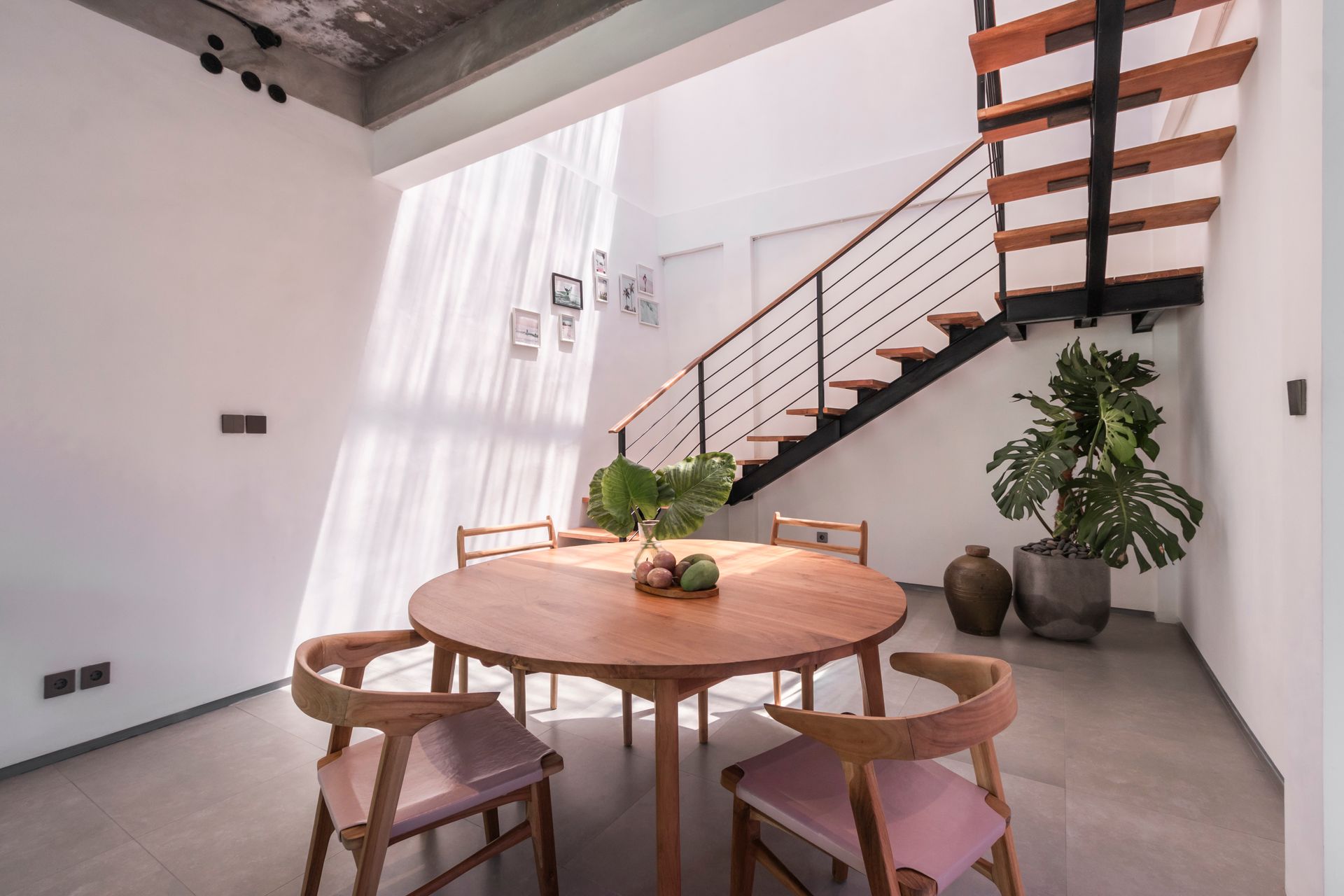 Dining area with wooden table, chairs, staircase, and potted plants. Bright sunlight streams through the window.