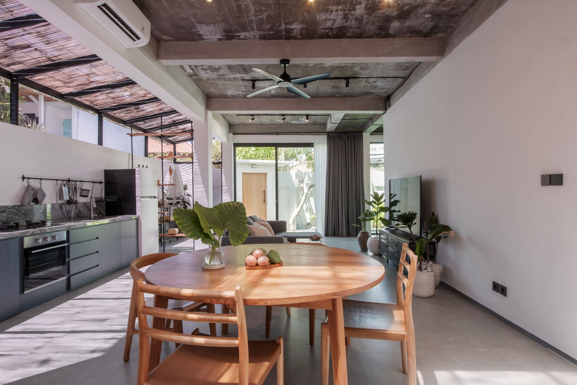 Open-plan living space with kitchen, dining area, and outdoor view. Wooden furniture, gray cabinets, and concrete ceiling.