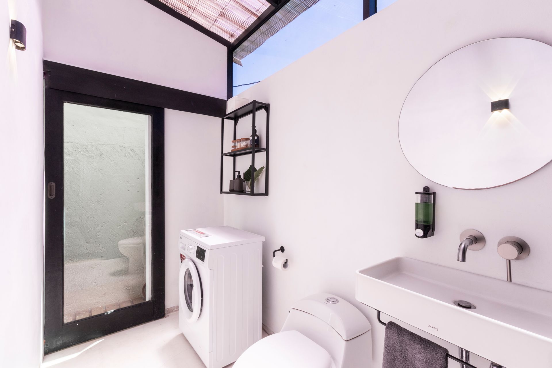Modern white bathroom with washing machine, sink, toilet, and shelving; natural light enters through a glass door.