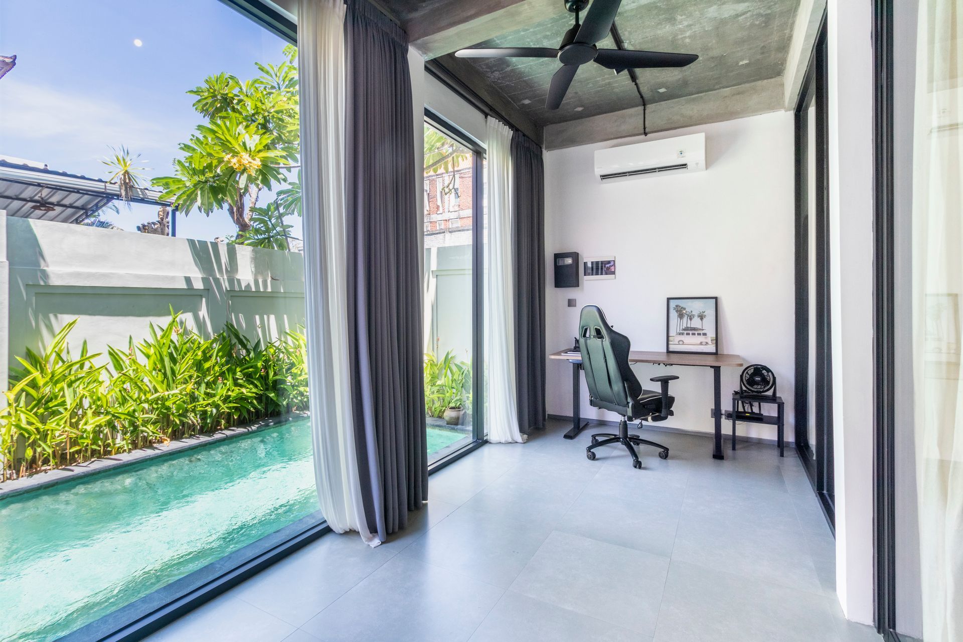 Office space with pool view, black desk and gaming chair, glass windows and gray curtains.