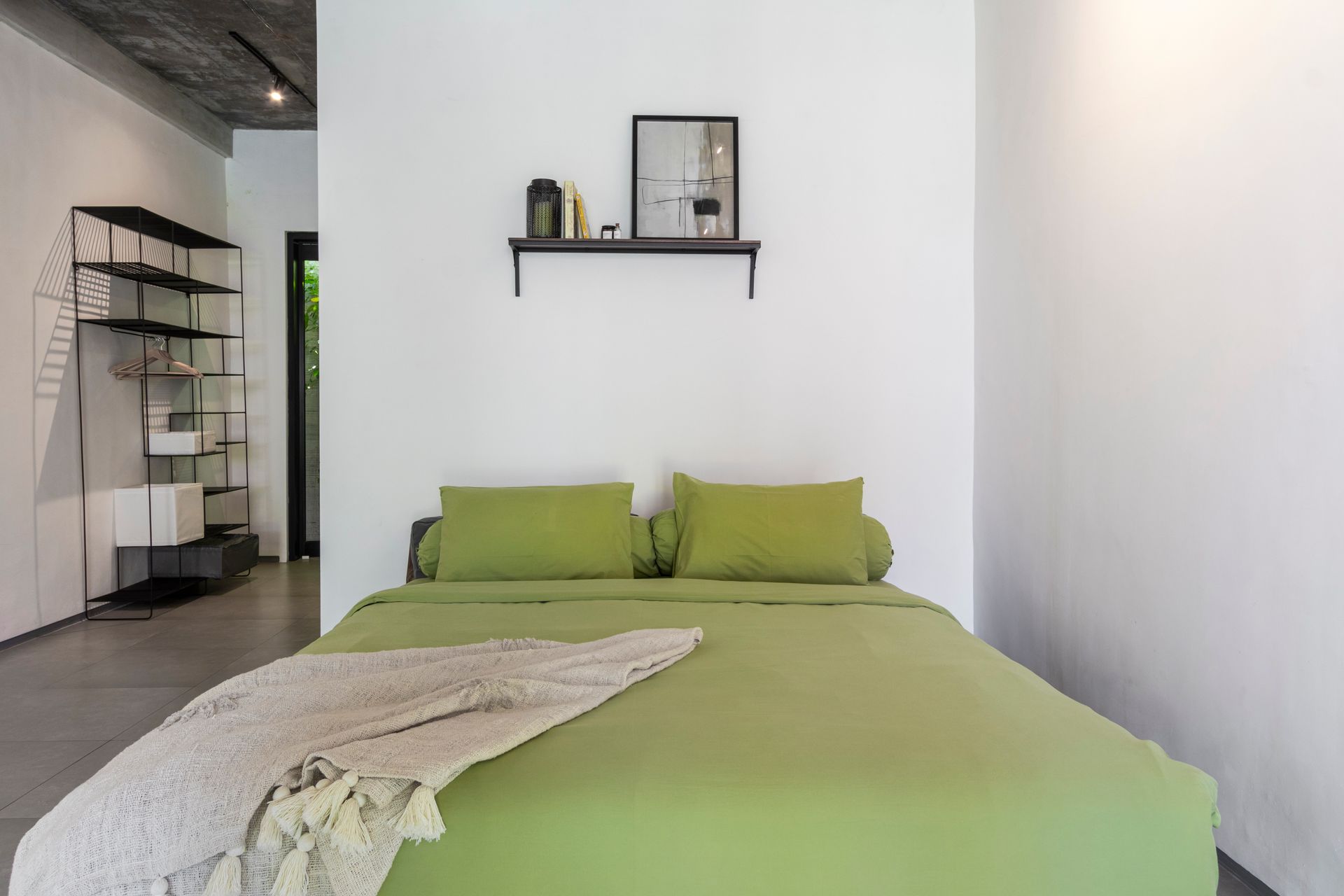 Green bed with pillows and a blanket in a modern, white-walled bedroom; shelf with decor.