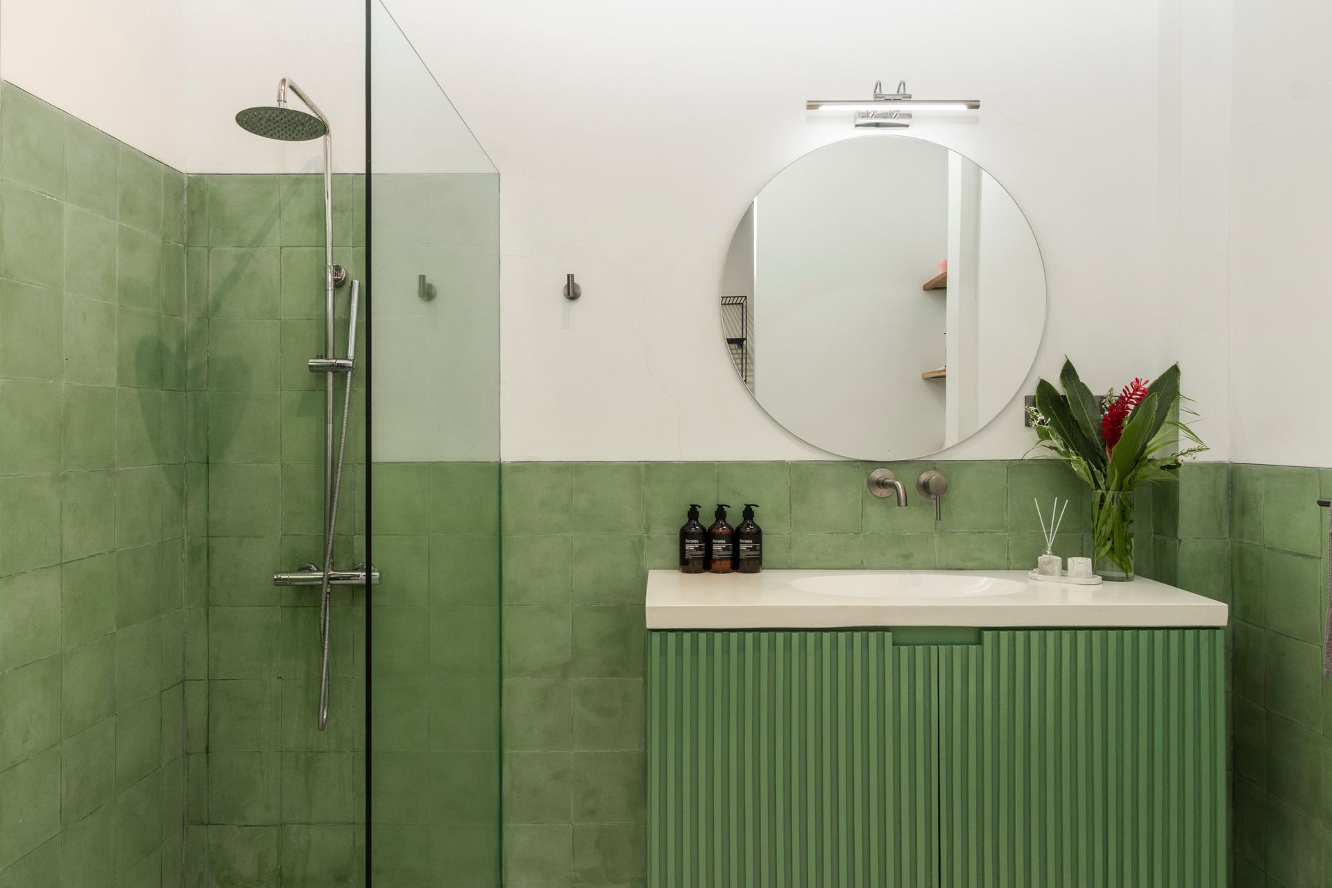 Green-tiled bathroom with shower, vanity, and round mirror. White walls. Vase of flowers on the counter.