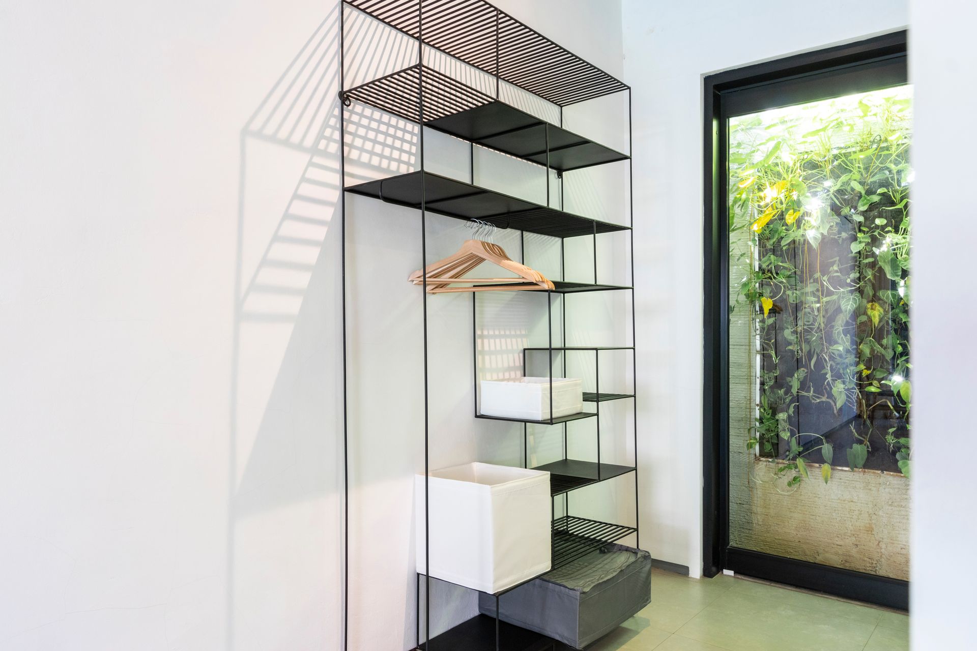 Black metal shelving unit against a white wall; holds white boxes, hangers, and sits next to a glass door with plants.