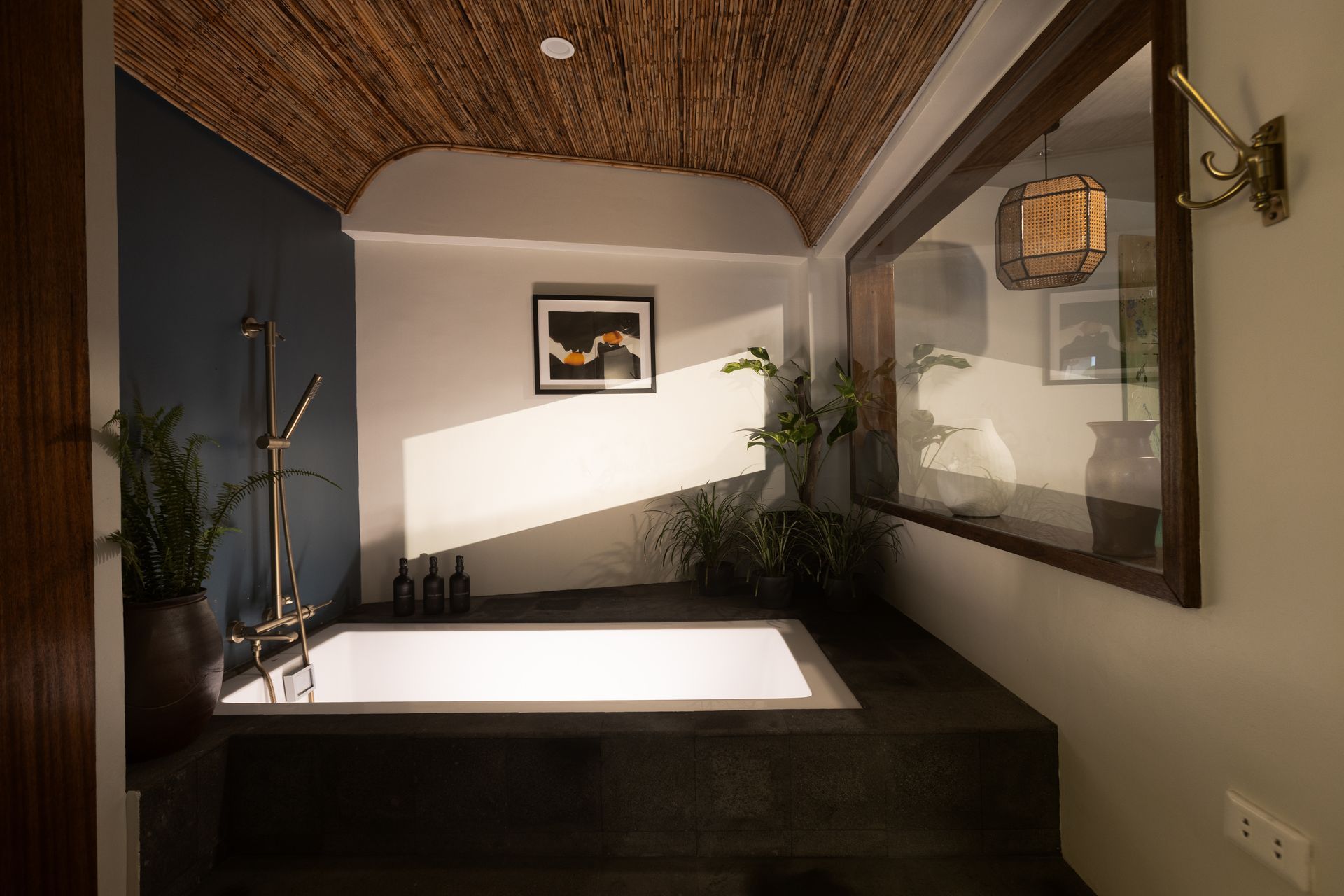 Bathroom with a sunken tub, dark blue and white walls, natural woven ceiling, plants, and a window.