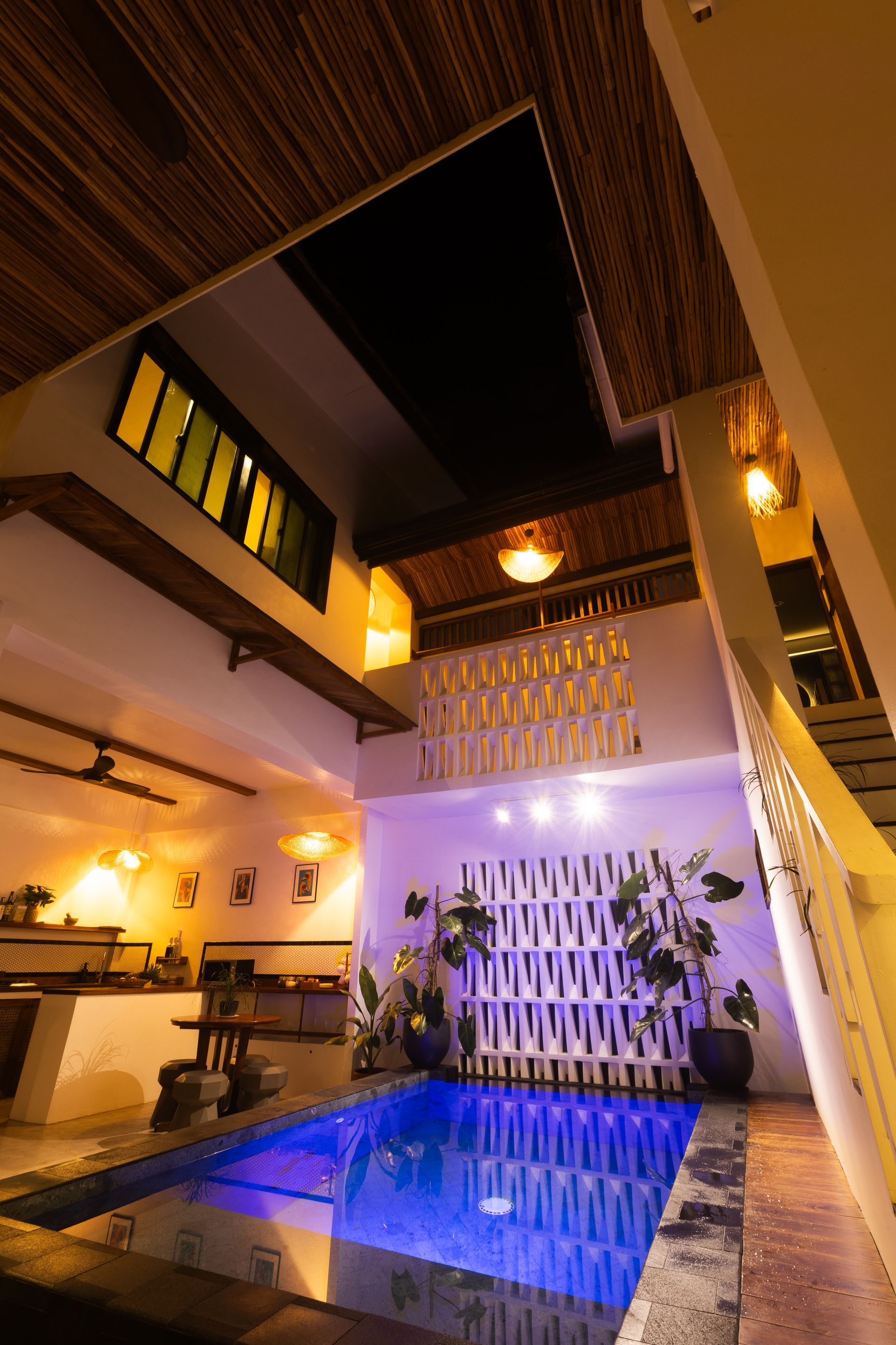 Indoor swimming pool with white walls, plants, and open ceiling. Blue water illuminated with lights.