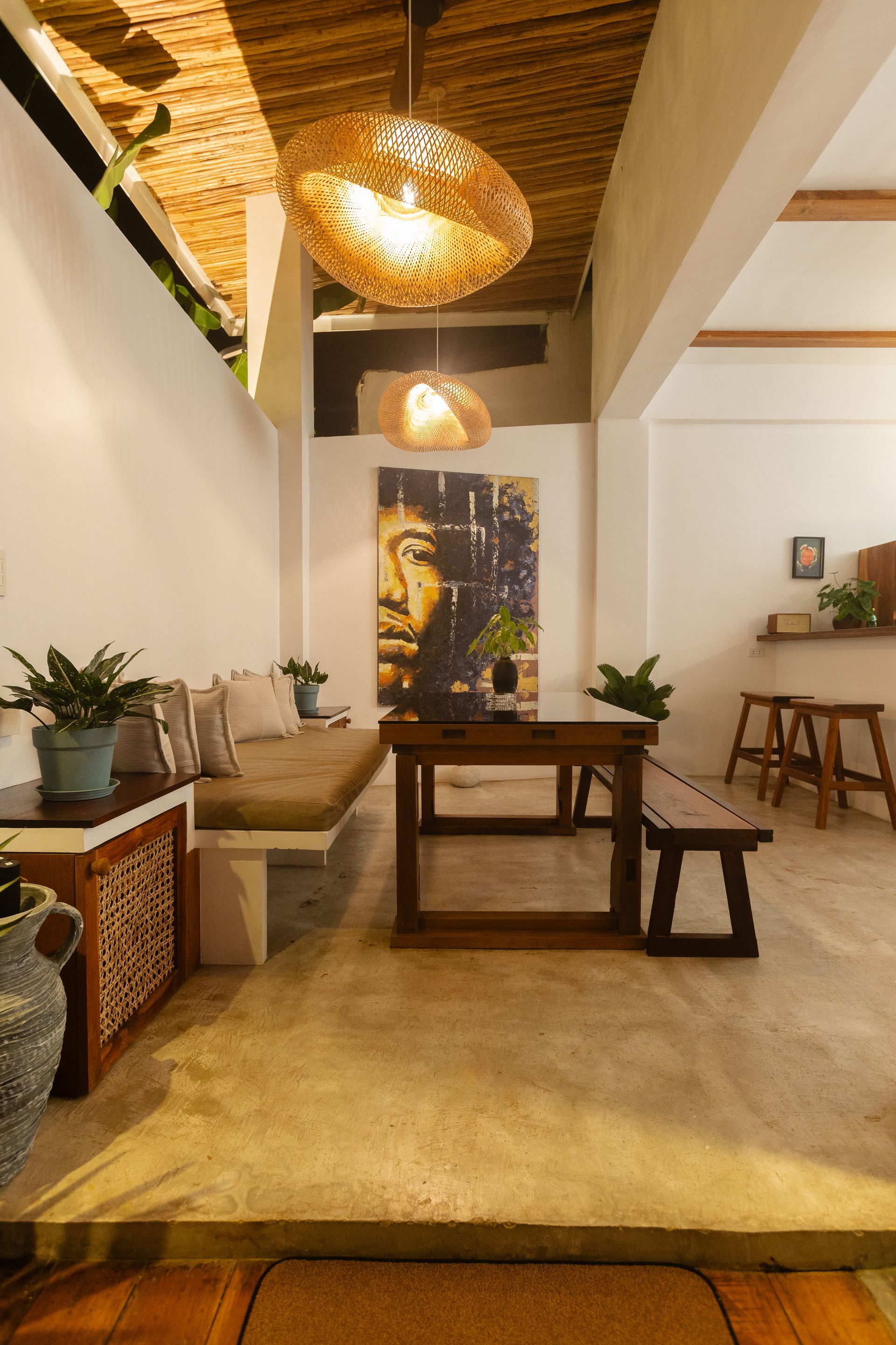 Interior with a dining area: table, bench, and a sofa with plants, under decorative pendant lights.