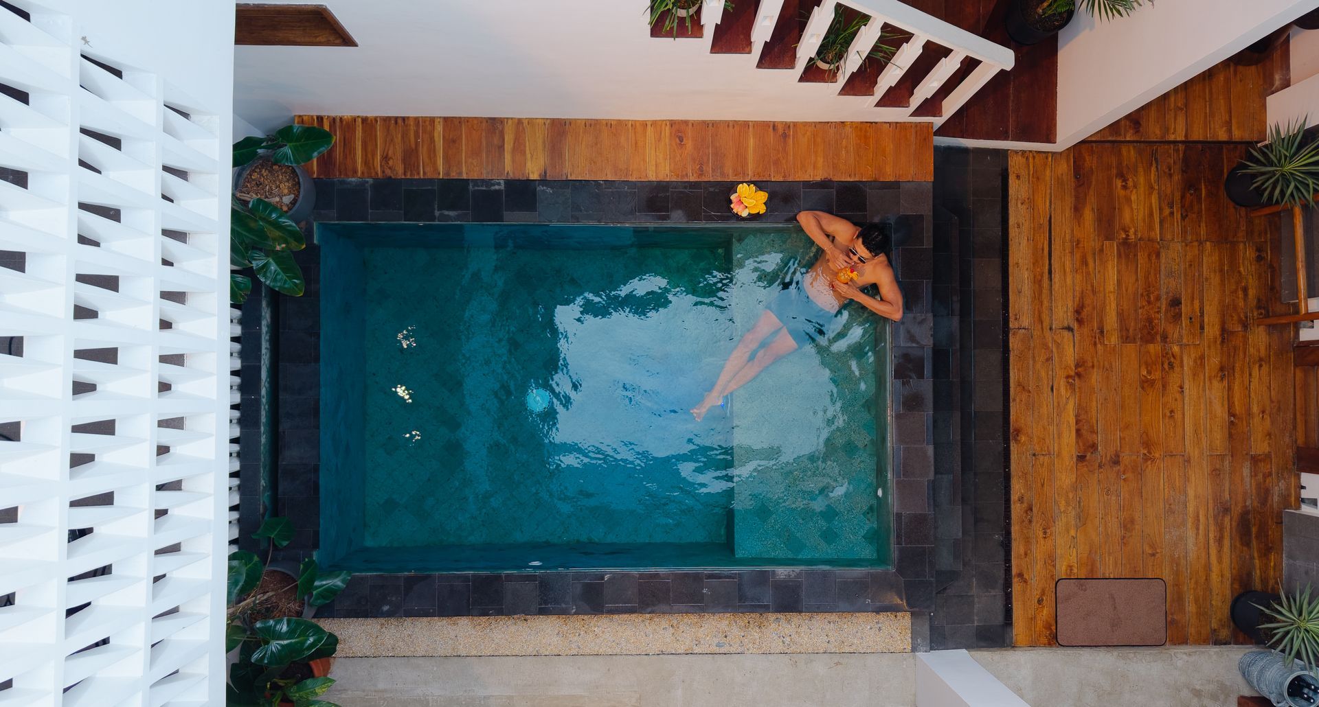 Person floating in a small, turquoise pool. Brown wood surrounds the pool and stairs above. Yellow flower floats nearby.