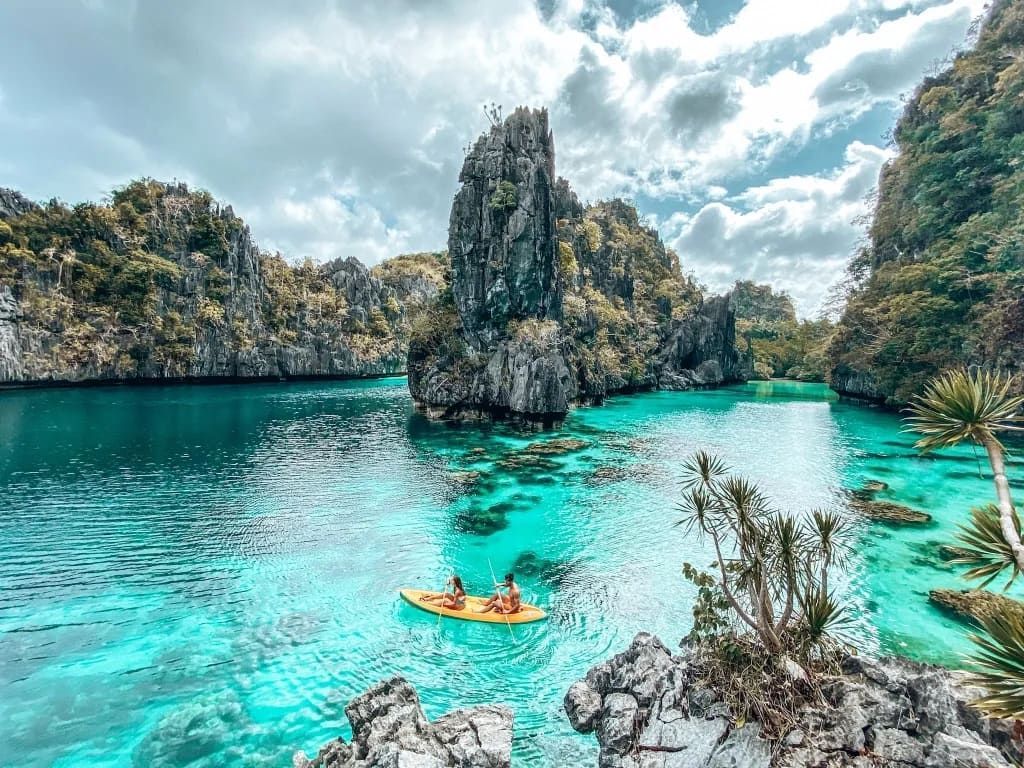 Kayakers in turquoise water surrounded by limestone cliffs and lush greenery under a cloudy sky.