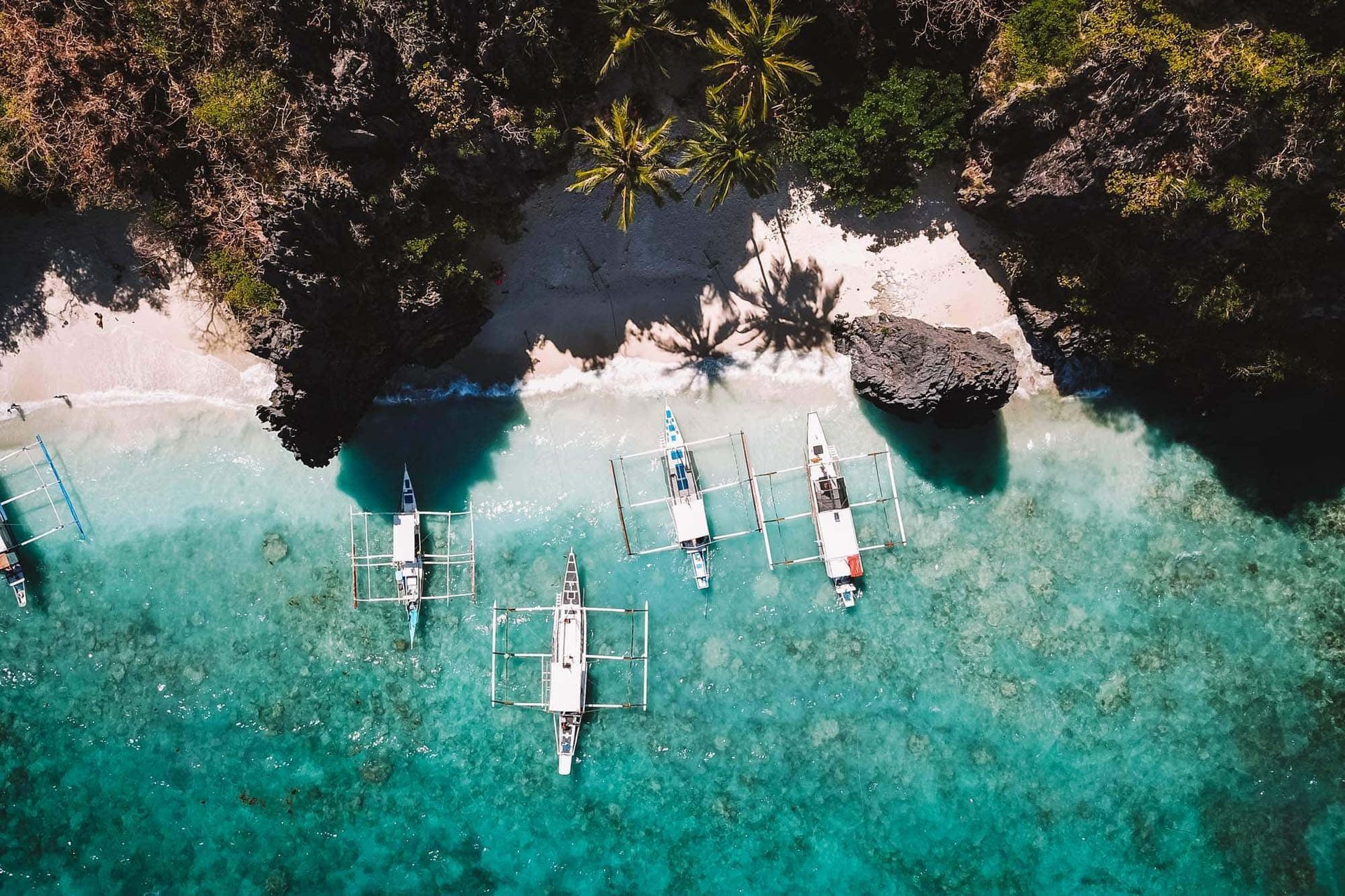 Aerial view of boats in turquoise water near a sandy beach with rocky cliffs and palm trees.