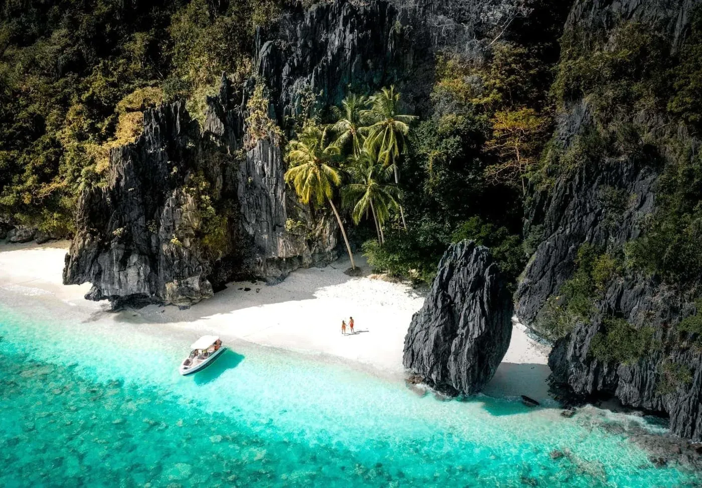 White sand beach with turquoise water, surrounded by dark cliffs and palm trees, boat at the shore.