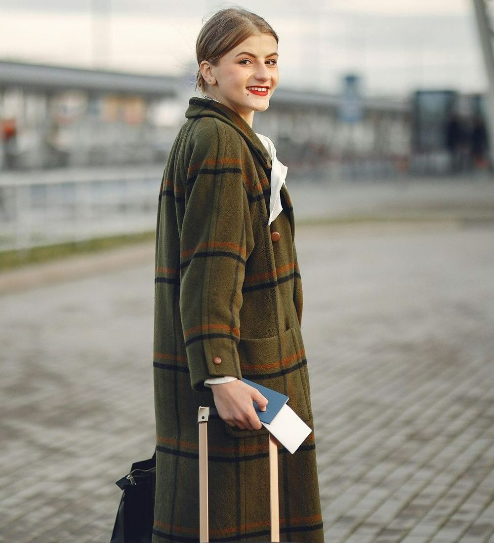 A woman in a green coat is holding a suitcase and a passport.