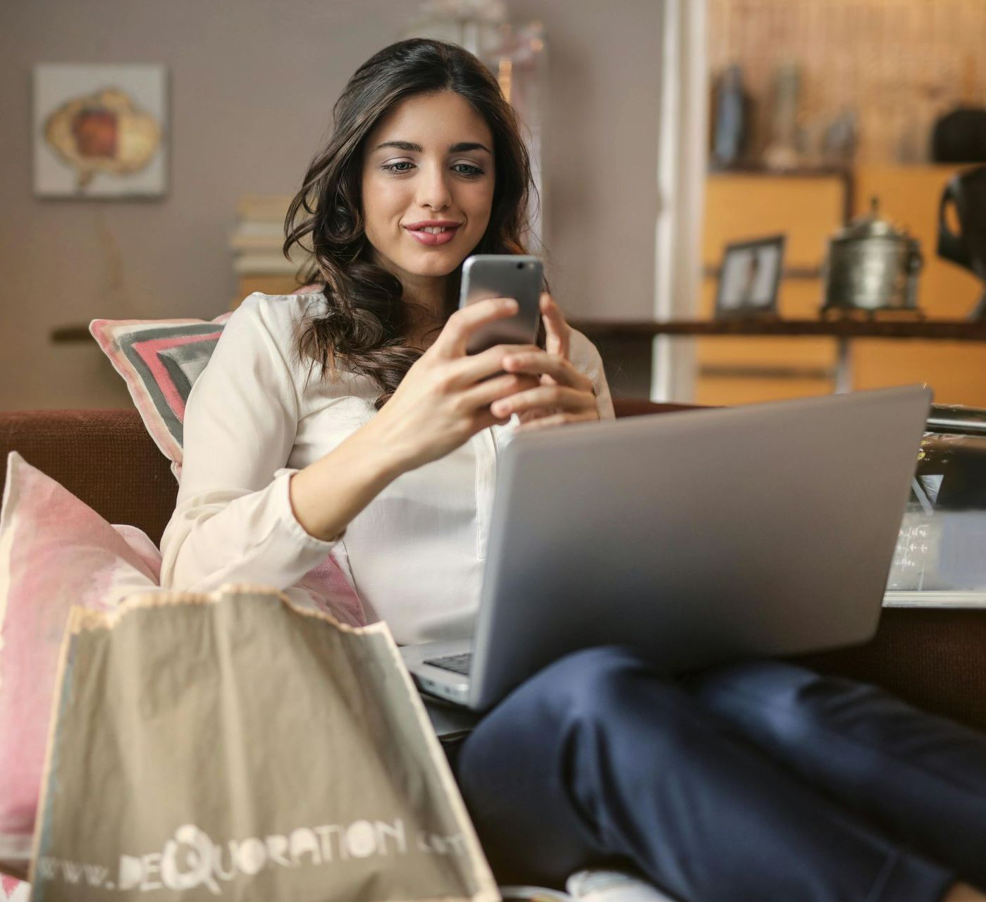 A woman is sitting on a couch using a laptop and a cell phone