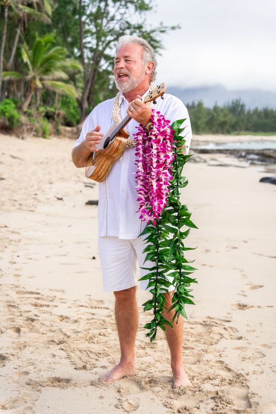 wedding officiant mark turansky stands playing the ukulele and holding lei for a hawaii beach wedding