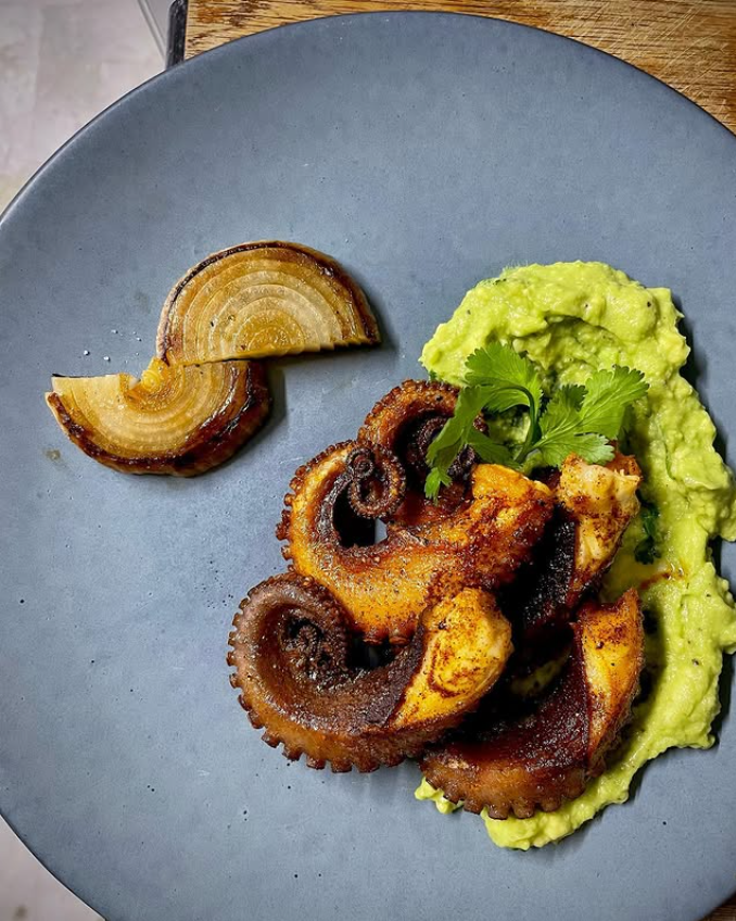 Un plato de comida con pulpo, guacamole y cebolla sobre una mesa.