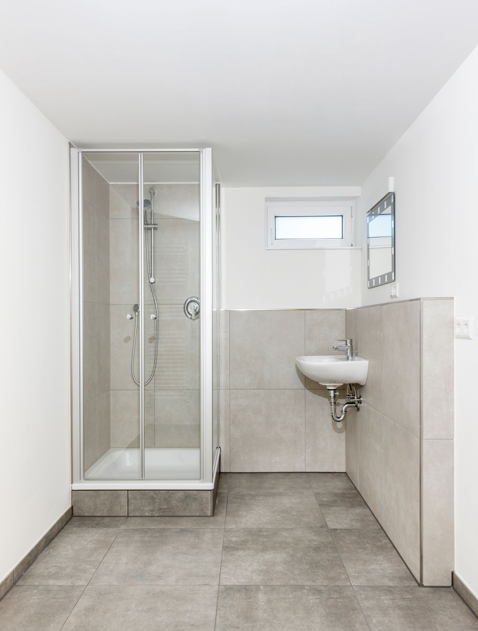 Modernes Badezimmer mit Dusche, Waschbecken, Fenster und grau gefliesten Böden und Wänden.