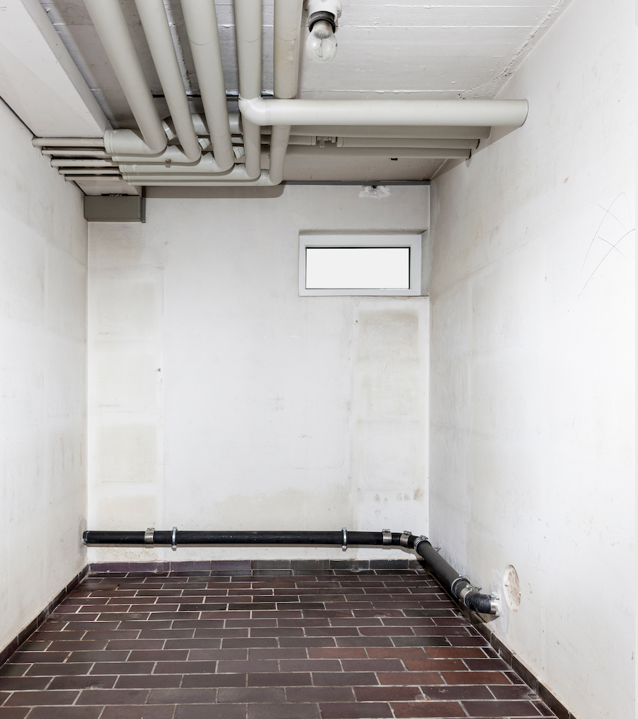 Innenansicht eines kleinen, weiß getünchten Zimmers mit Ziegelboden, Rohren an der Decke und einem kleinen Fenster.