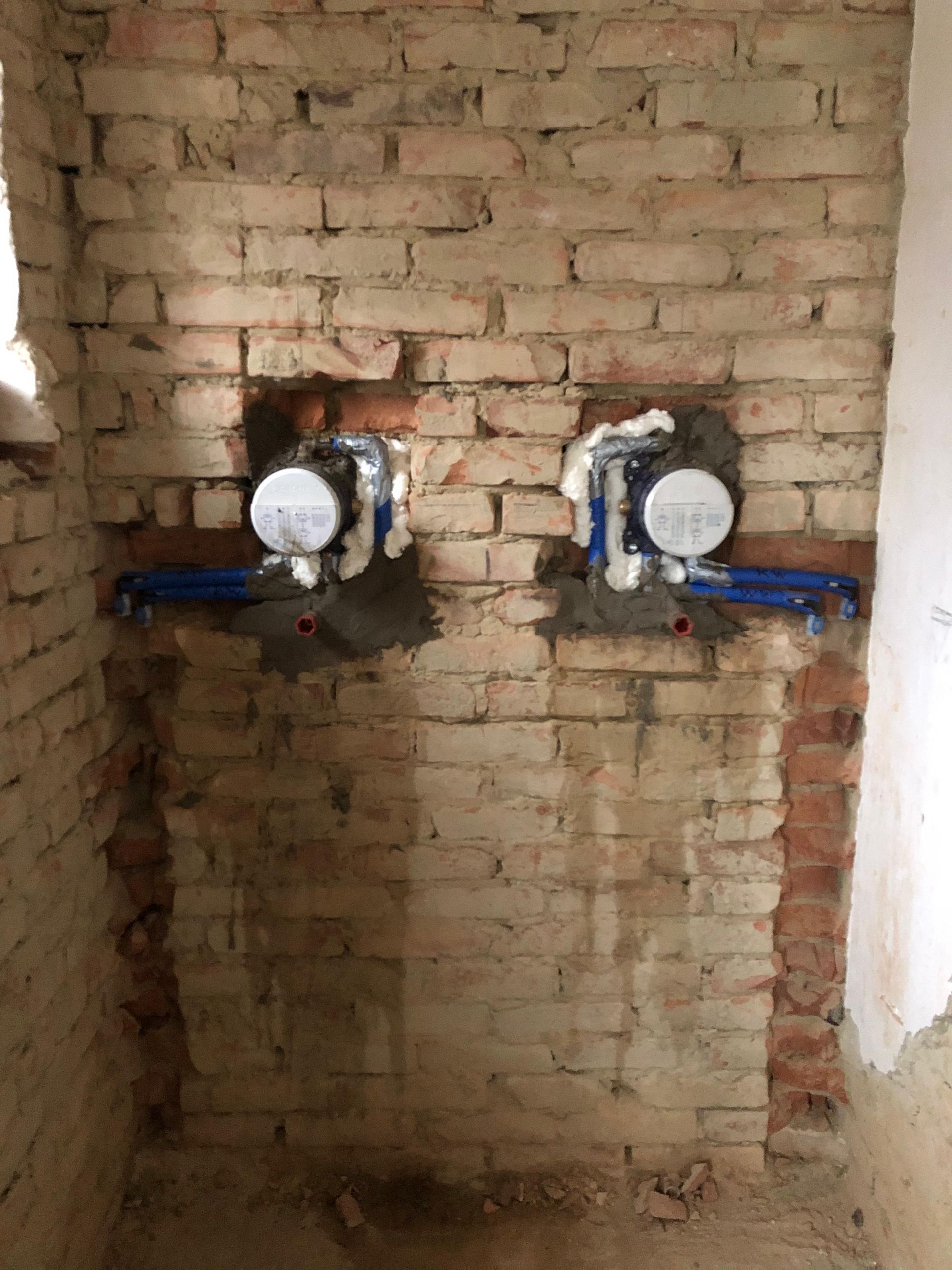 Zwei silberne Sanitärarmaturen sind in eine Ziegelwand mit blauen Rohren eingelassen und bereit für die Fliesenverlegung.