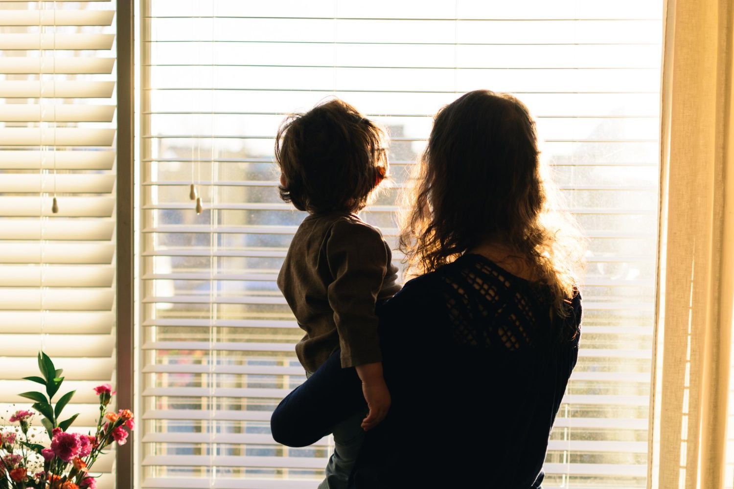 Person holding a child in front of a bright window with blinds