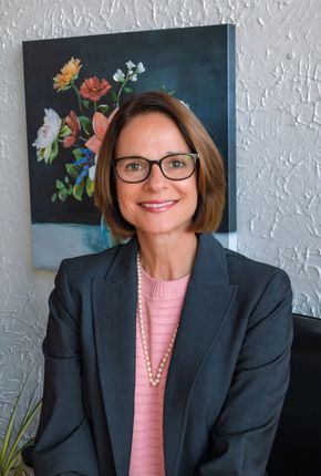 Woman in glasses and dark blazer standing before a floral painting and white textured wall.