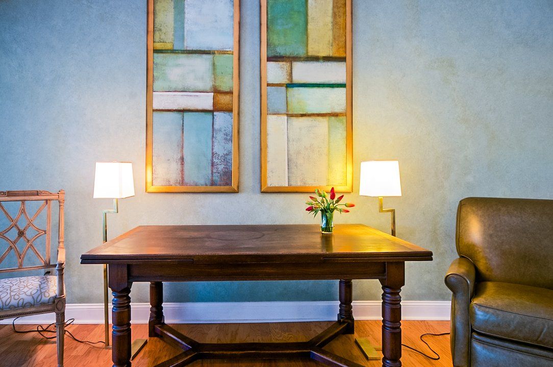 A living room with a table , chairs , lamps and two paintings on the wall.