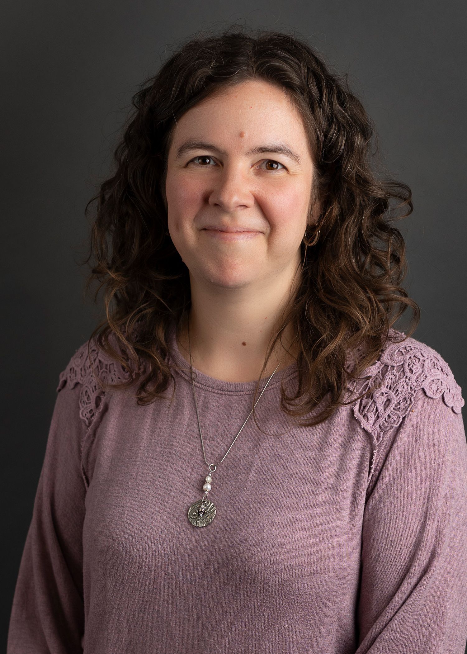 A woman with long brown hair and a necklace is smiling for the camera.