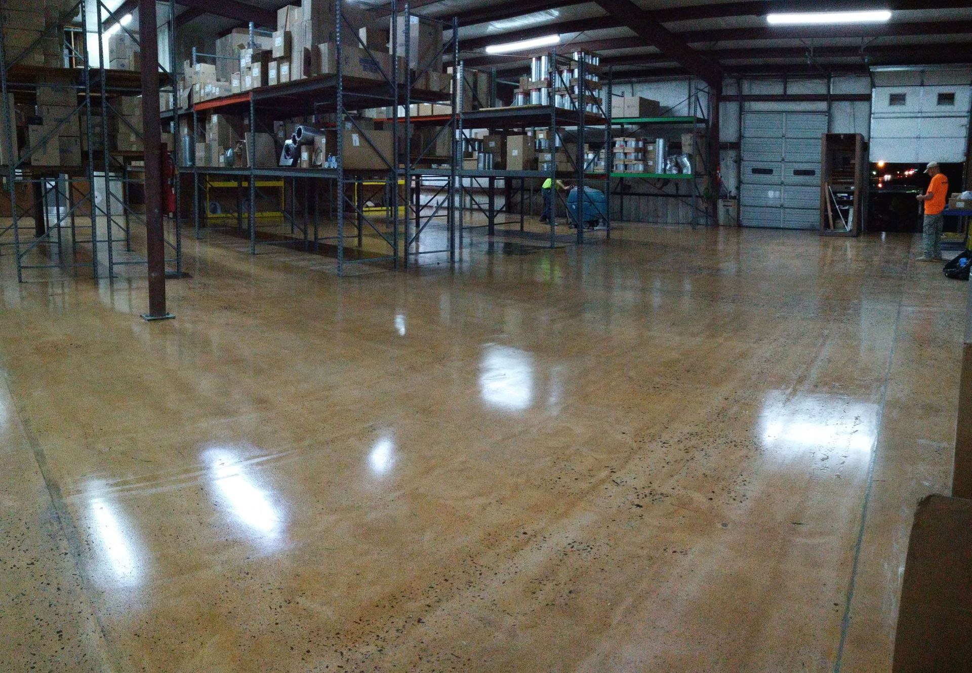 Polished concrete warehouse floor reflecting overhead lights. Shelving and boxes are in the background.