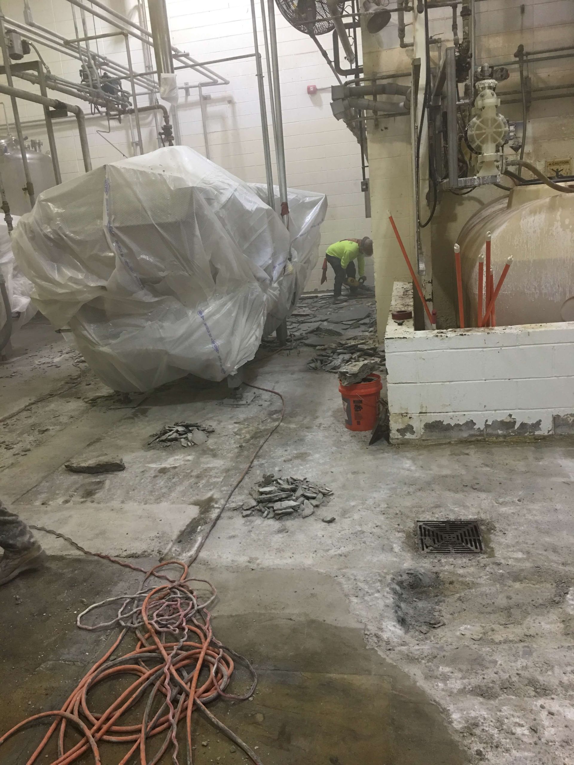 Construction site with workers removing concrete floor. Large covered equipment visible, industrial setting.