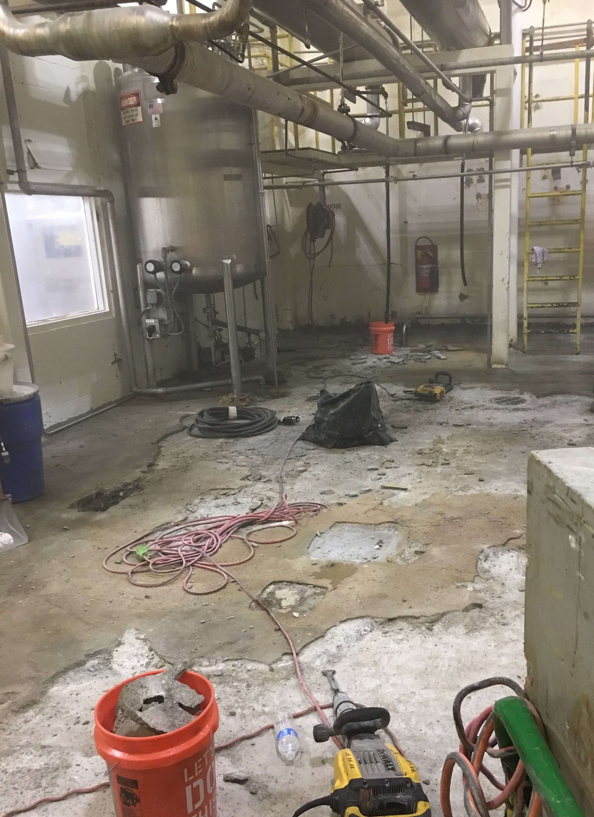 Industrial interior with damaged concrete floor, construction tools, and piping overhead.