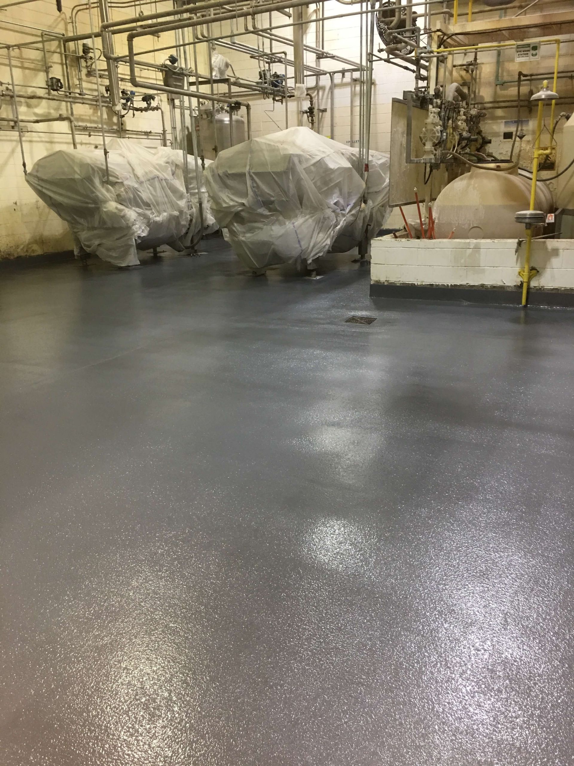 Gray industrial floor with equipment covered in plastic; pipes overhead.