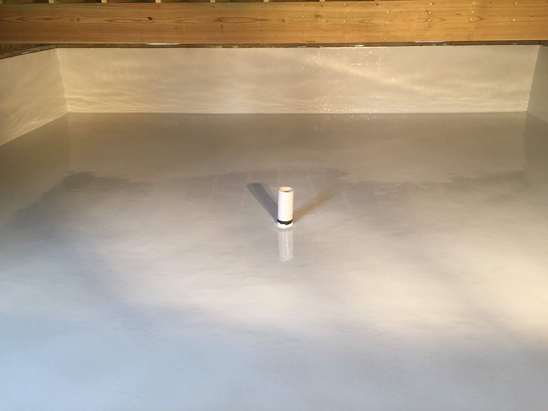 Empty, light grey-painted rectangular room with a white vertical pipe centered on the floor, wooden beam overhead.