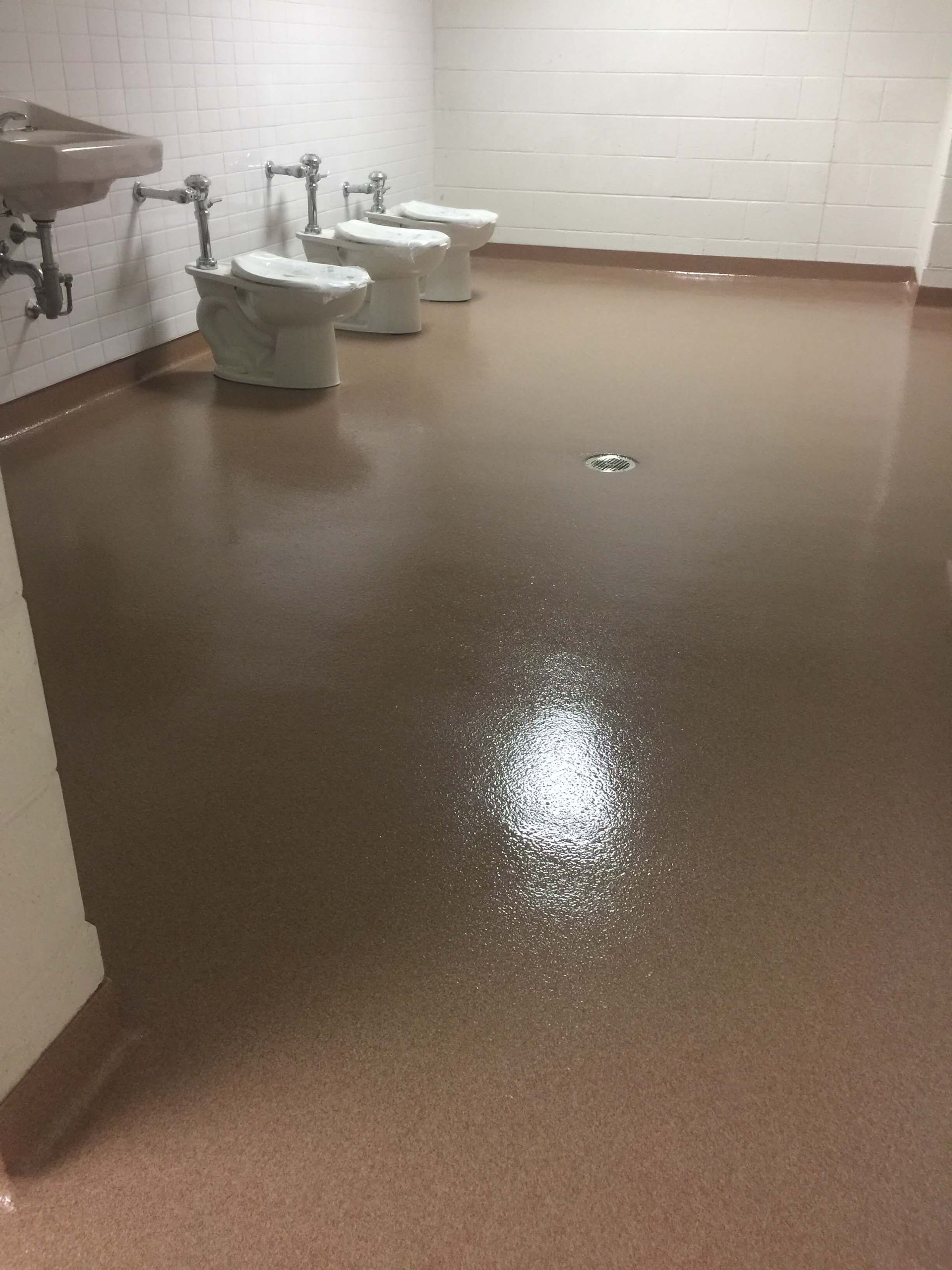 Public restroom with brown floor, white toilets, and sinks. Water is visible on the floor.