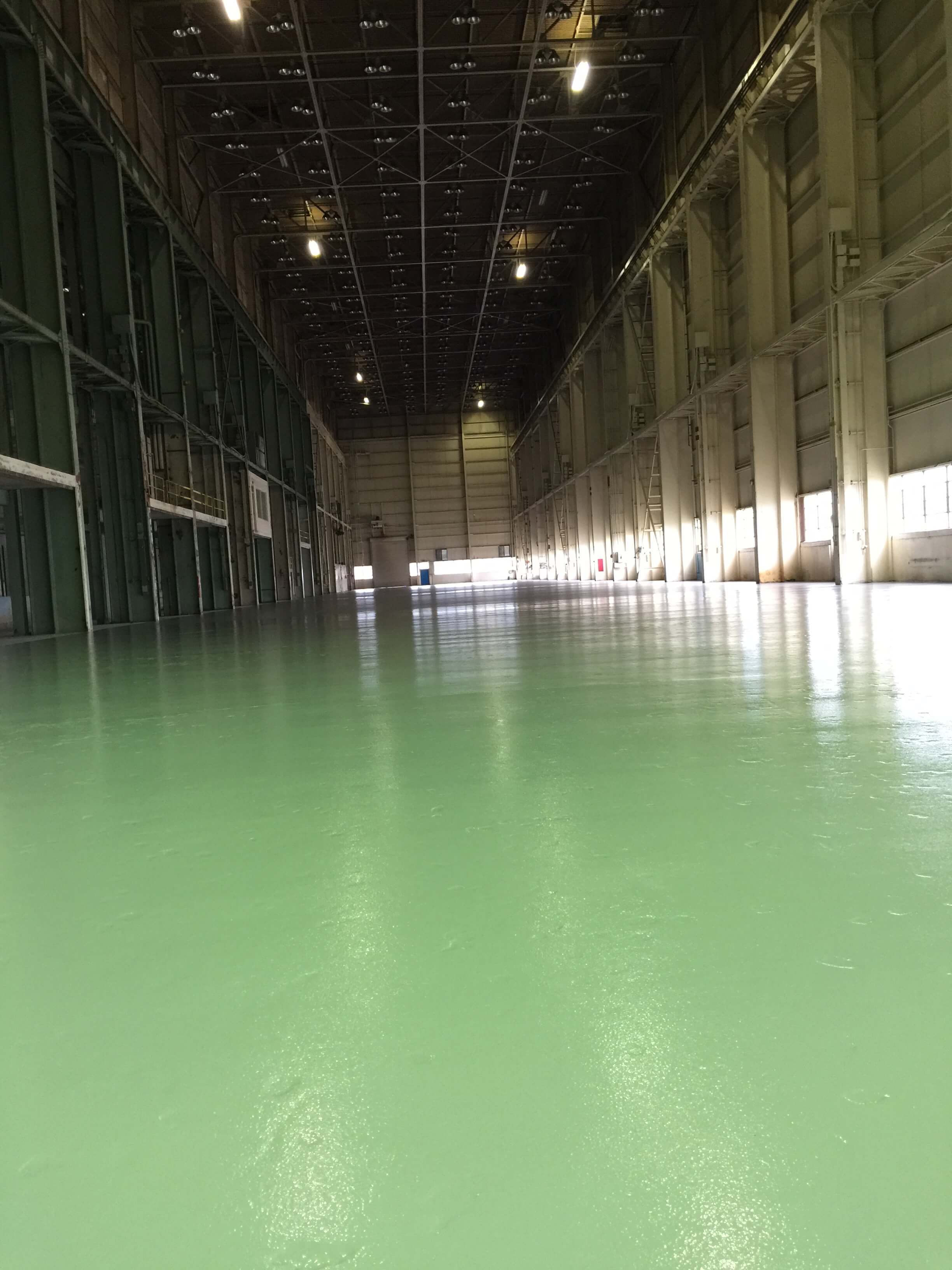 Interior of a large warehouse with a smooth, green floor reflecting light; open doors at the end.
