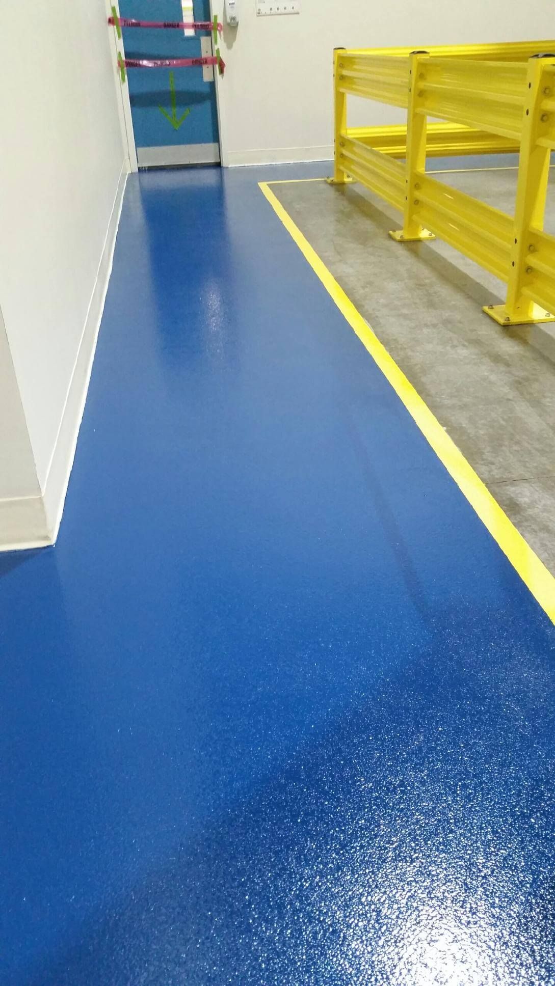Blue epoxy floor with yellow line, next to white wall. Yellow safety rails in the right.