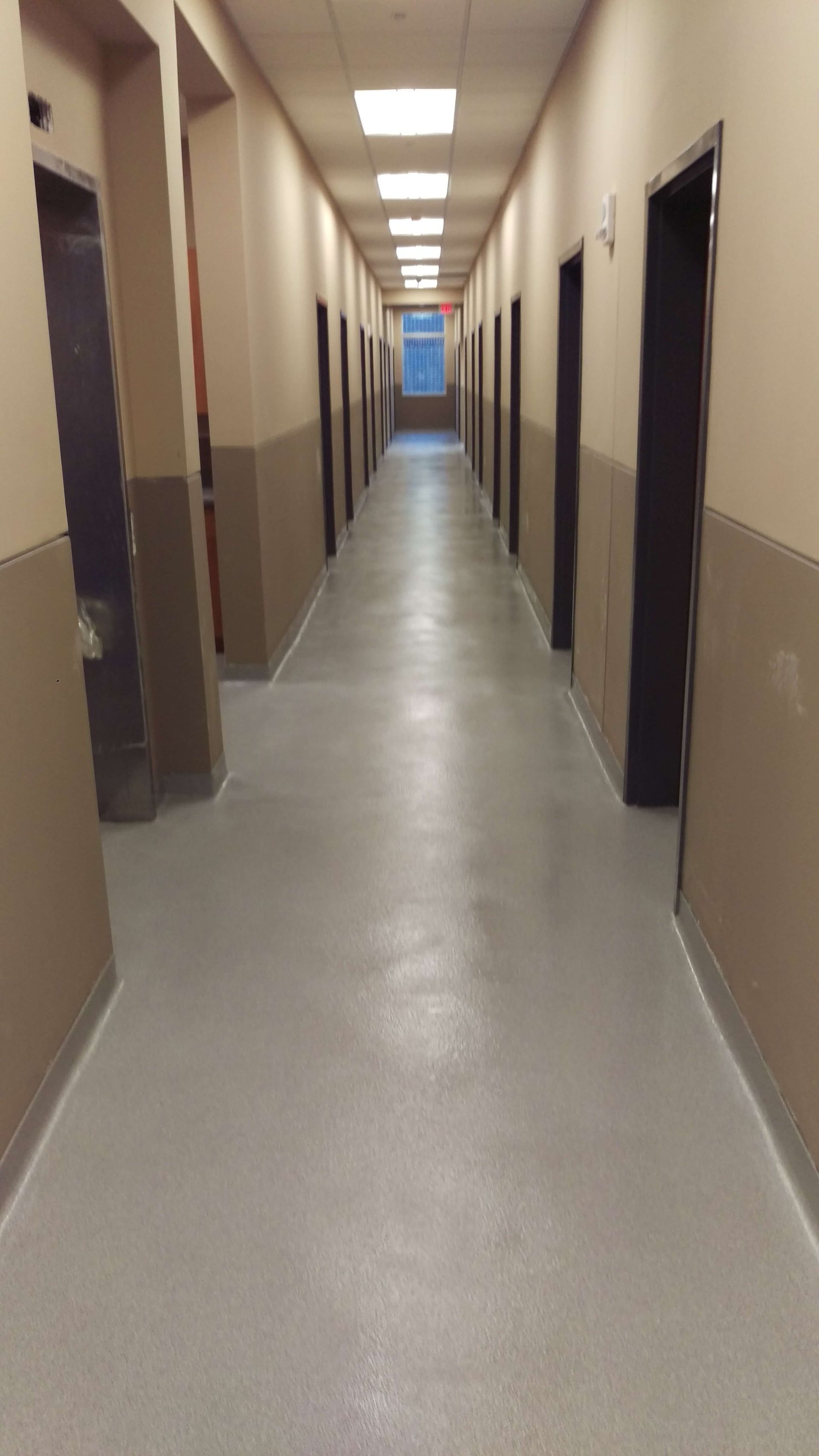 Long hallway with closed doors, beige walls, gray floor, and overhead lighting.
