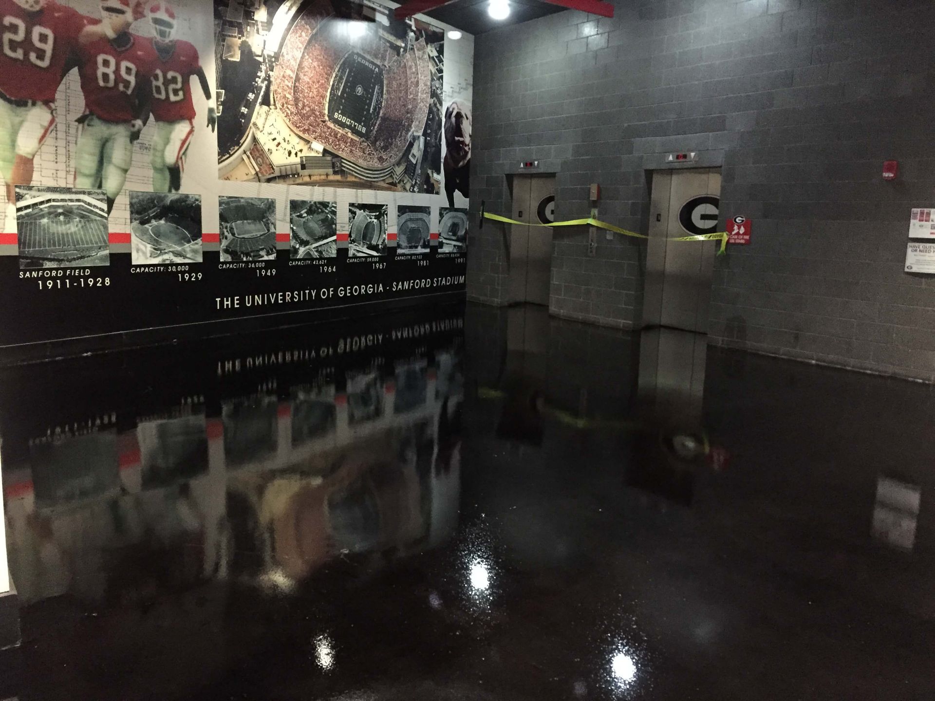 Flooded interior near University of Georgia elevators, with yellow caution tape. Black water reflects walls, displays.