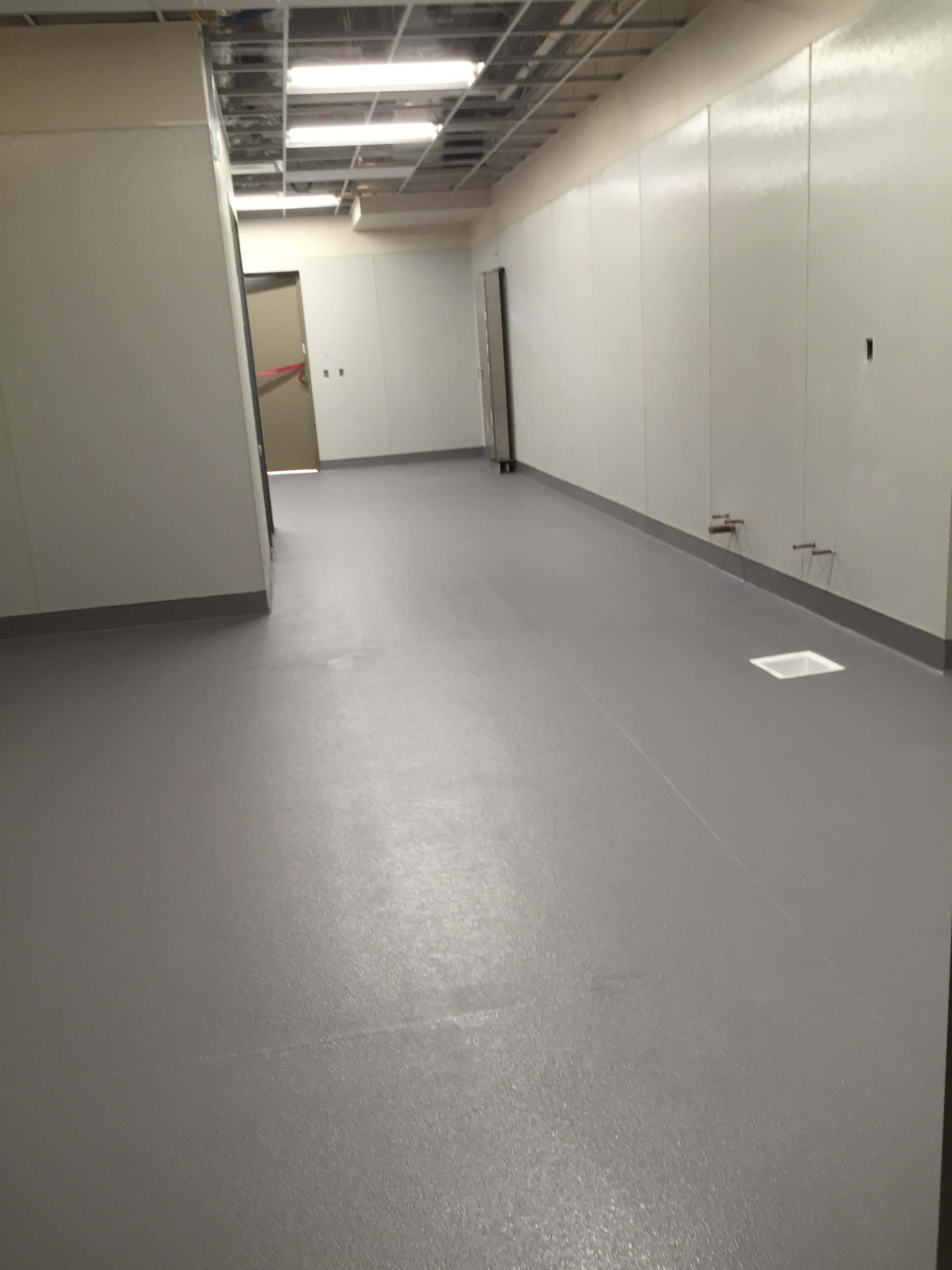 Empty room with light gray flooring and white walls. A door is visible at the end of the hallway.