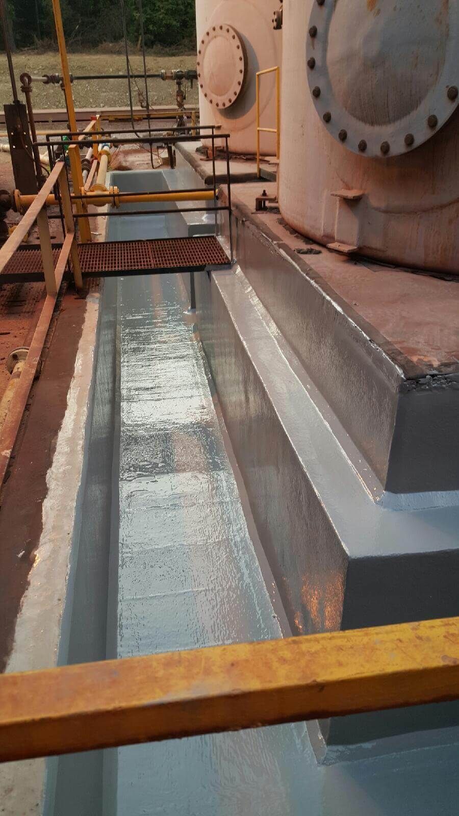 A narrow concrete trough with a light-gray coating, next to large industrial tanks and yellow metal walkways.